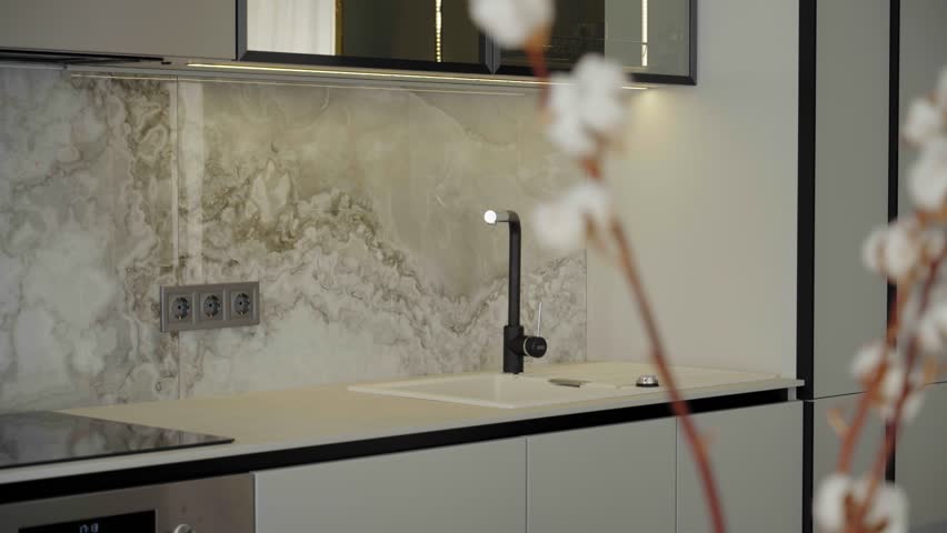 Modern bright kitchen interior with a marble backsplash and black faucet, photographed through softly blurred cotton branches in the foreground