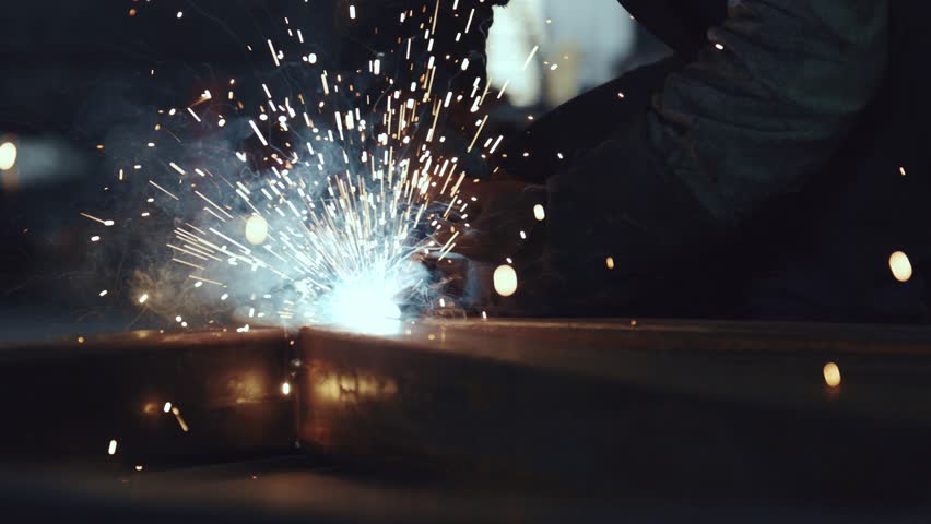Close up of a welder's hands connecting steel parts, creating bright sparks and smoke in a dark industrial workshop. The professional is using equipment for metalwork and construction