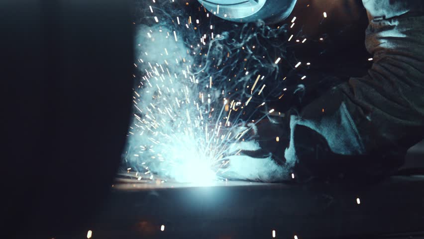 Professional welder in a protective mask and gloves welding metal parts in a dark workshop, creating bright sparks and smoke during the industrial manufacturing process in a factory