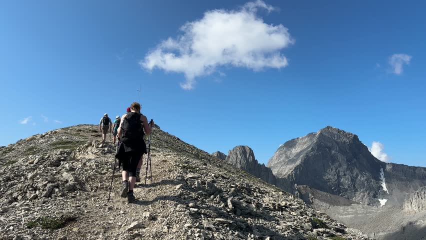A Caucasian female hiker climbing Tent Ridge in Kananaskis, Alberta, using hiking poles, towering Rocky Mountain peaks rising beneath a vivid blue sky