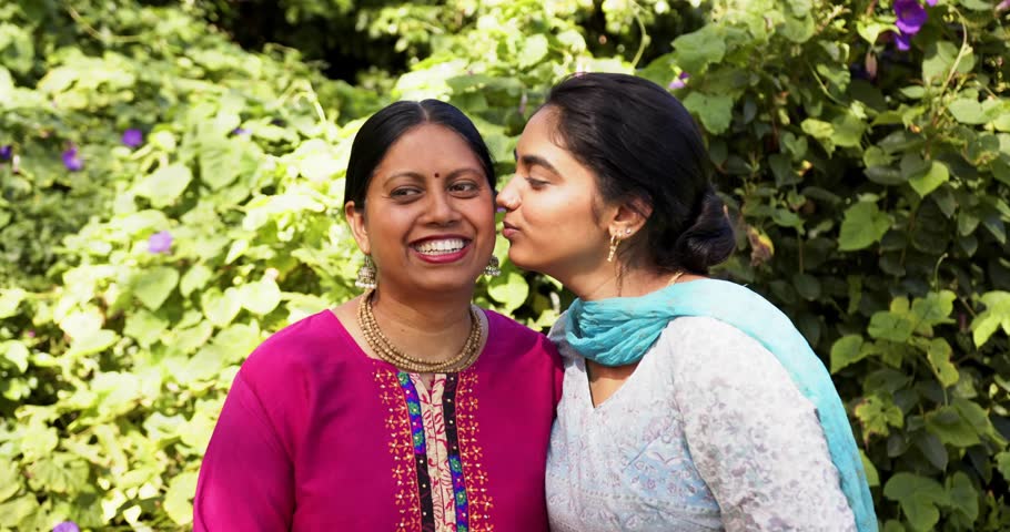 Daughter leaning in kissing mother by vine wall, sharing joy, generating social badges around faces. Foliage, garden, kurta, dupatta, embroidery, jewelry, affection
