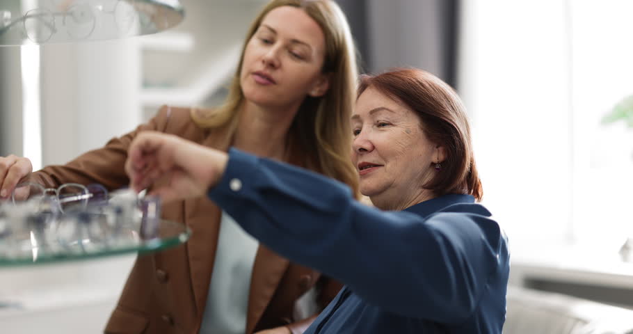 Close up two happy women, senior mother and young adult daughter standing near display shelf in optical store and trying on eyeglasses together. Vision health for younger and older, eyewear selection