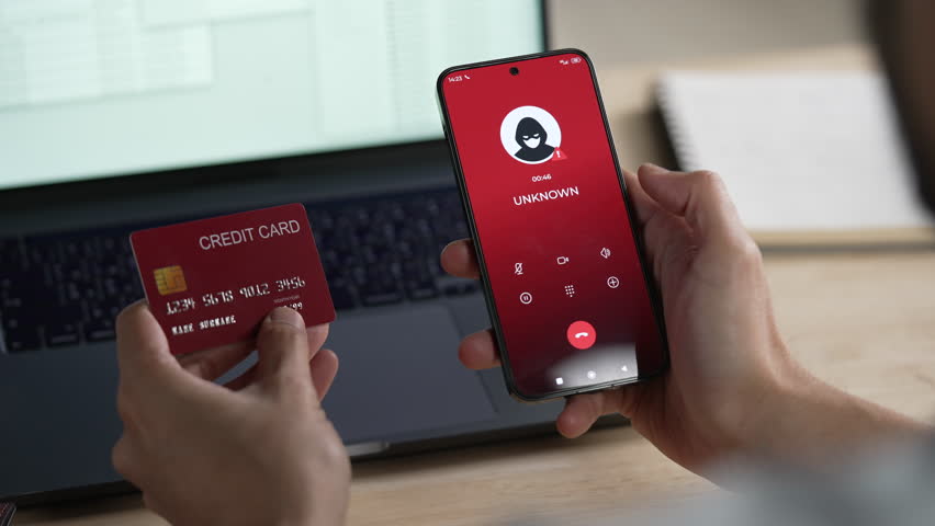 A credit card user giving the credit card number to a scammer while holding a phone showing a scammer on the line