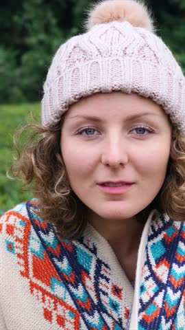 Beautiful young woman with curly hair and blue eyes wearing a knitted pom pom hat and a colorful patterned sweater, gently smiling at the camera outdoors with a green natural background