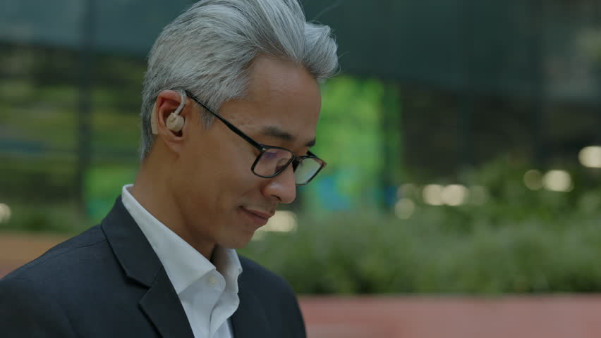 Closeup portrait of mature Asian businessman in glasses looking down at a laptop out of frame with focused expression. Concentrated corporate worker sitting outdoors near modern office buildings
