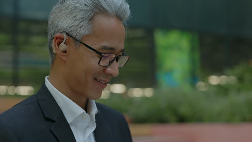 Closeup portrait of mature Asian businessman in glasses looking down at a laptop out of frame with focused expression. Concentrated corporate worker sitting outdoors near modern office buildings