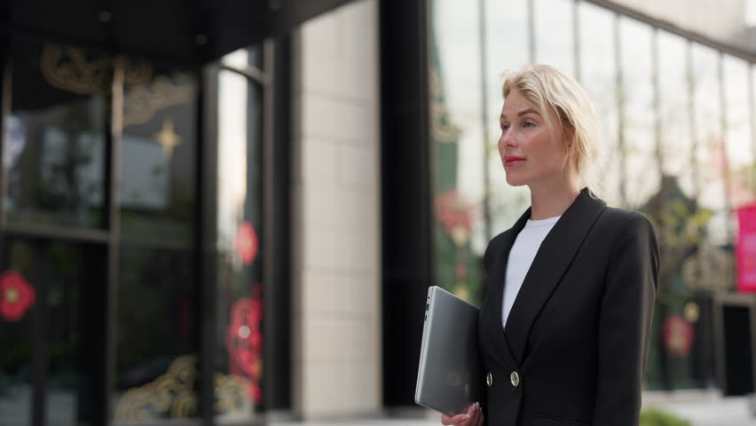 Portrait of focused businesswoman with laptop walks past commercial buildings slow motion. Young blonde entrepreneur in black jacket with notebook computer strolls on city street