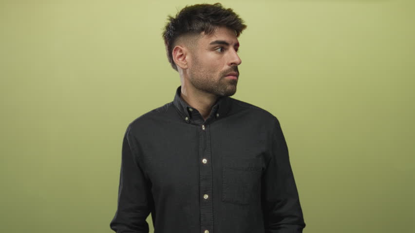 Young hesitant hispanic man buttoning shirt collar with both hands against light green wall in studio; fear anxiety dread worry.