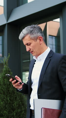 Man going outdoor with mobile phone in his hands. Middle aged businessman walking using smartphone outdoors in city near office building. Slow motion.