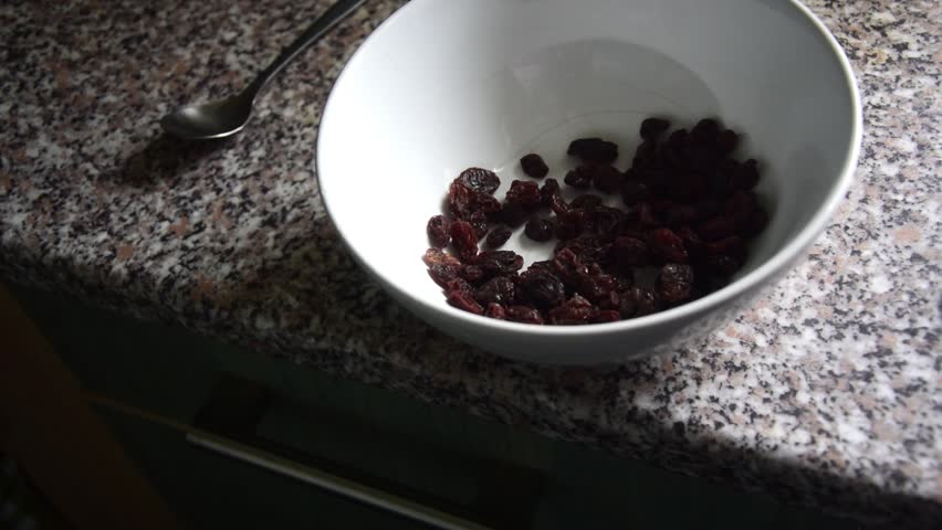 Slow motion close shot of a white bowl on a kitchen worktop, with someone pouring raisins into it