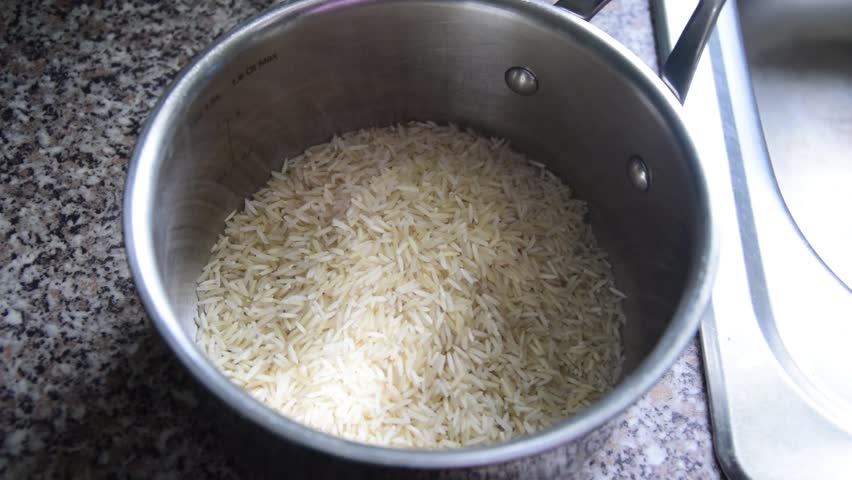 Slow motion close shot of a stainless steel saucepan on a kitchen worktop next to a sink, with someone pouring white rice into it