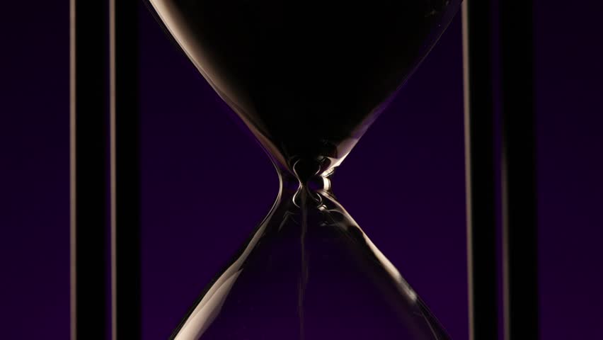 fine grains of sand passing through the narrow waist of a transparent hourglass in front of purple background