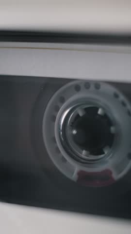 Vertical, extreme close-up of a transparent audio cassette tape playing. The white plastic supply reel spins as the dark magnetic tape unwinds inside the vintage player, showing its mechanism.