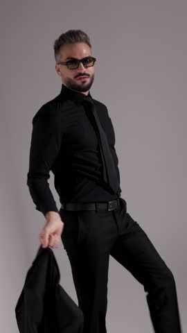 Cool man in sunglasses and all black outfit is putting his jacket on his shoulder and walking away on grey background.