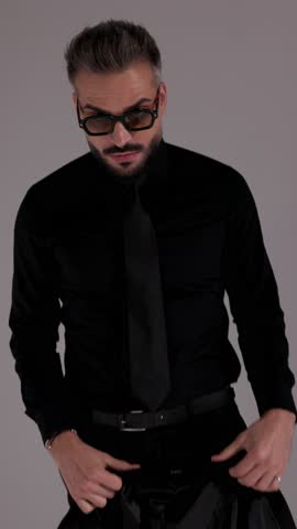 Cool man in sunglasses and all black outfit is putting on his jacket and walking away on grey studio background.