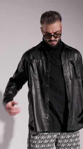 Cool man in sunglasses and leather jacket is fixing his jacket, putting hands in pockets, fixing his glasses, and touching his chin on white background.