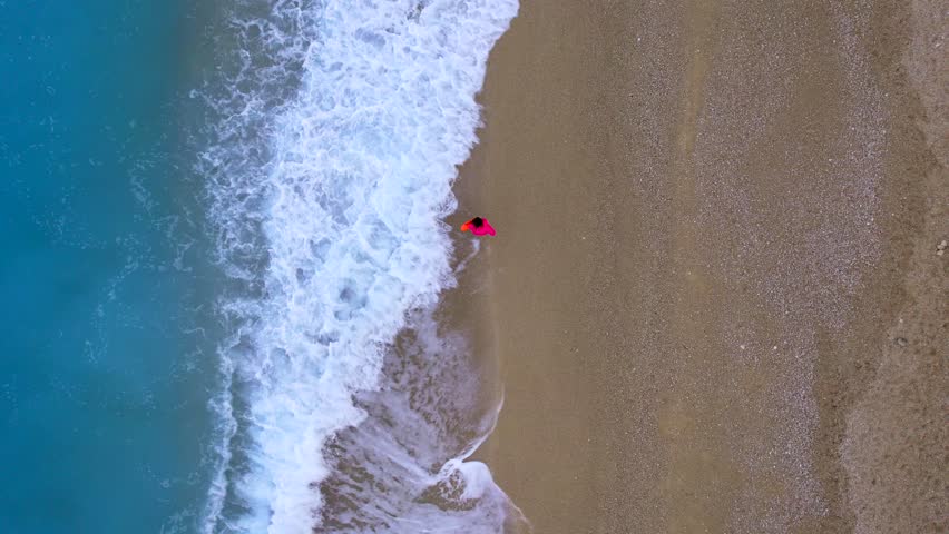 Aerial view of woman in pink coat walking by the shoreline on a peaceful sandy beach as sea waves roll in beside her