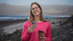 Woman in pink sweater smiles holding smartphone and coffee cup at the seaside; digital connection serenity. - Powered by Shutterstock - Get 15% off with code: PIKWIZARD15