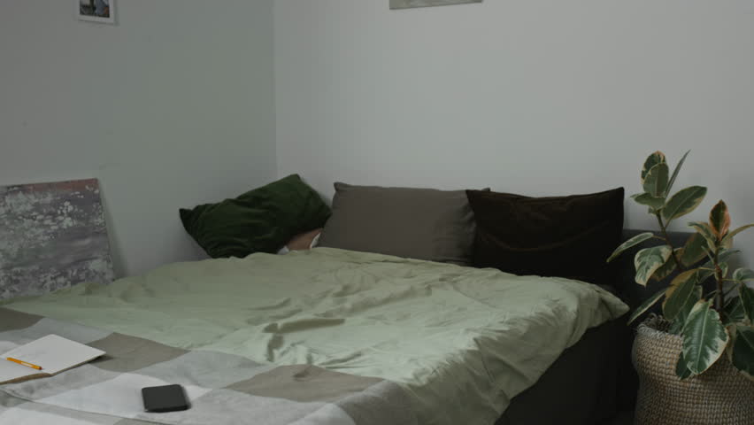 Young woman with alopecia taking off her blond wig and lying down in bed with her laptop to work from home