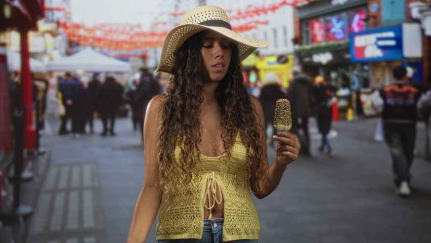 Young woman holding a chocolate coated icecream on a stick points finger up toward camera while wearing a sunhat and crochet top in a crowded street market; confidence attitude.
