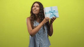 Woman holds a white gift box with blue ribbon and shakes it while listening in a studio; anticipation discovery. - Powered by Shutterstock - Get 15% off with code: PIKWIZARD15