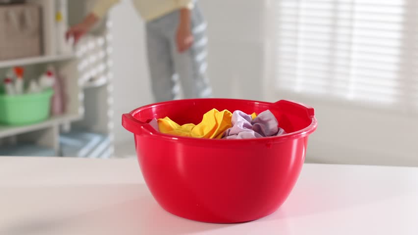 Woman pouring detergent into plastic basin with clothes at table indoors, closeup. Camera moving forward