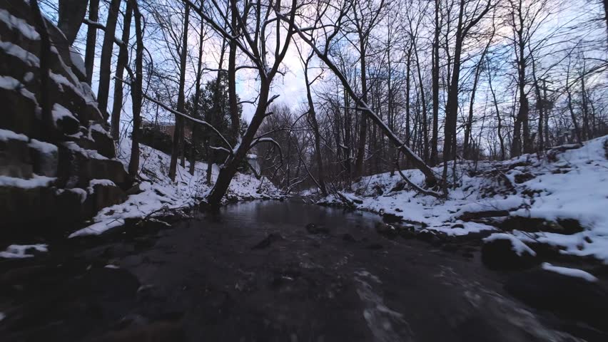 Smooth FPV dive into a narrow cold winter creek path surrounded by snow, ice, and leafless forest.