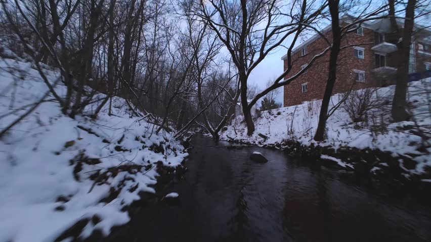 Dynamic FPV chase following a snowy creek bed surrounded by bare winter trees and icy water flow.