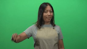 Woman showing peace sign hand and smiling in a green studio while wearing beige apron and gray tshirt, short black hair; playful confidence. - Powered by Shutterstock - Get 15% off with code: PIKWIZARD15