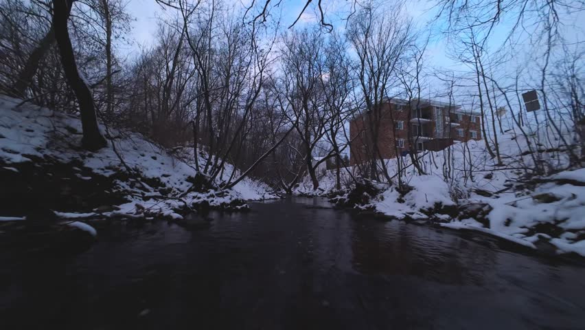 Dynamic FPV tracking along a shallow winter creek with icy water and snow-covered slopes on both sides.