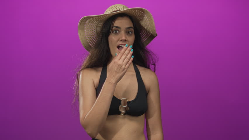 Woman wearing black bikini top and straw hat with hand covering mouth laughing in purple studio; joy.
