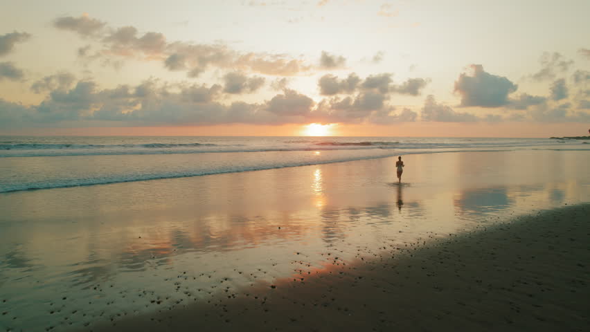 Woman walking into ocean during vibrant sunset with reflections on wet beach