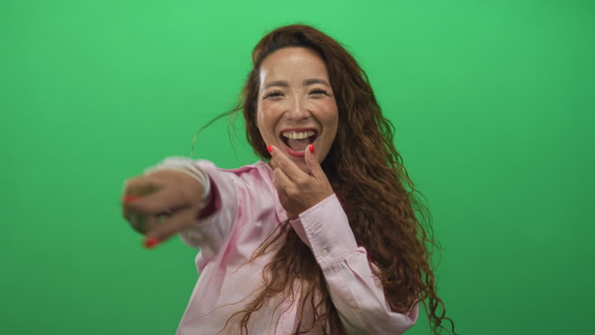 Woman in pink shirt smiling with hands on cheeks and open mouth laughing in green studio; surprise joy warmth.