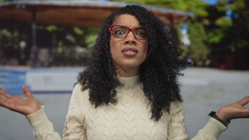 African american woman with curly hair and red glasses shrugs hands in forest setting under wooden shelter; confusion frustration perplexity uncertainty.