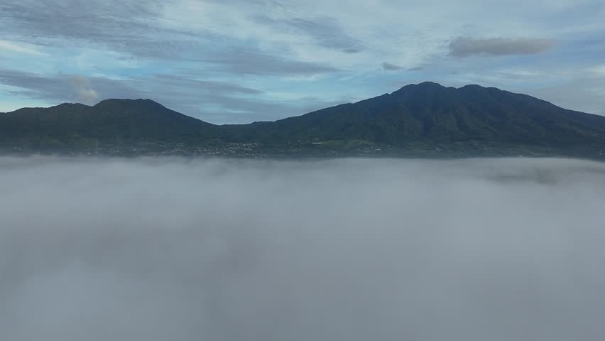 A calm mountain range rises above a blanket of soft white fog, framed by a blue sky with gentle clouds, creating a peaceful and refreshing natural scene