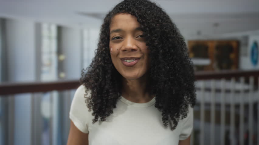 Woman with curly hair wearing white t shirt smiling and cupping hand to mouth for call gesture in hotel; excitement.