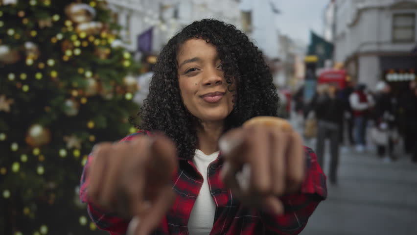 Woman in red plaid shirt points finger at camera on busy city street by christmas tree; friendly invitation.
