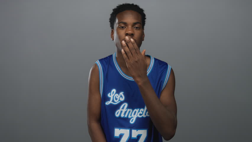 African american man in a blue basketball jersey with hand to lips blowing a kiss in a grey studio setting; affection.