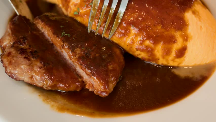 Close-up of fork slicing juicy hamburg steak beside omelette rice, rich brown sauce, warm lighting