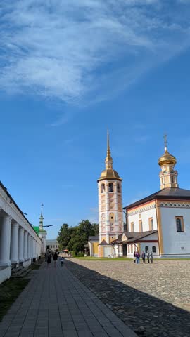 View of Suzdal old town. Golden domed cathedral under blue sky in center, sunlit cobblestone path leading toward ornate orthodox tower. Bird flying. Touristic place, vertical motion