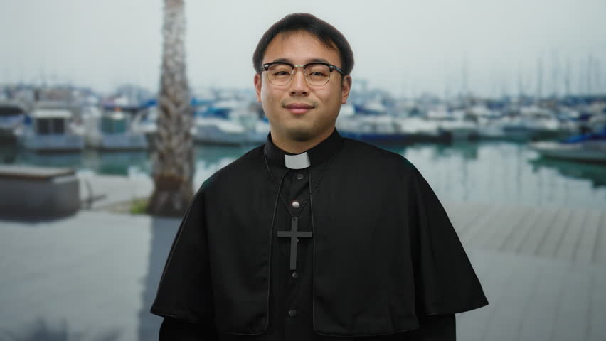 Man in religious robe standing in front of a marina with boats in the background, showcasing a serene expression by the seaside.