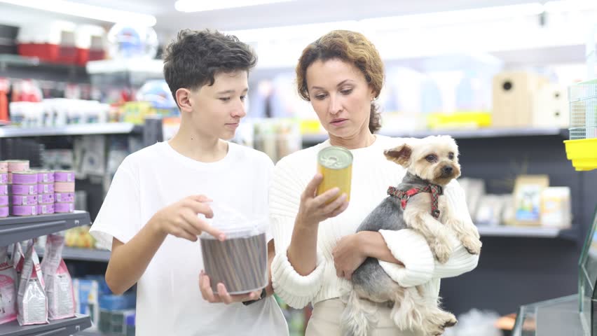 Caring teenage boy with mother visiting pet store with cute little Yorkshire terrier to buy dog food, reading ingredients on treat packages and discussing choice