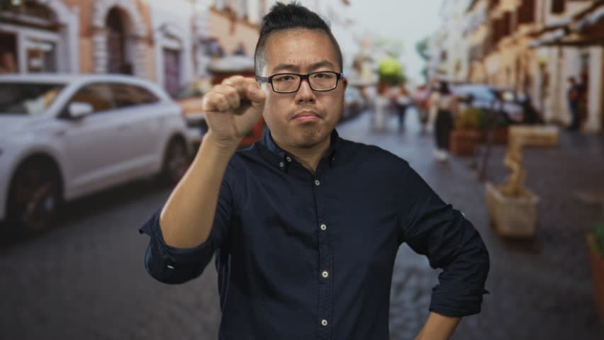 Young chinese man wearing glasses and navy shirt points finger forward on city street near parked car; quiet concentration.