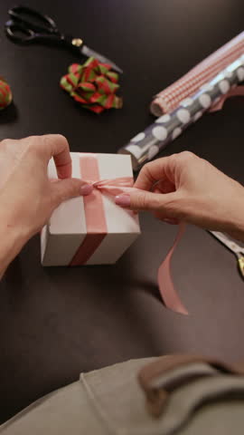 Vertical POV shot of hands of unrecognizable gift shop worker tying ribbon on box at working table while packing order