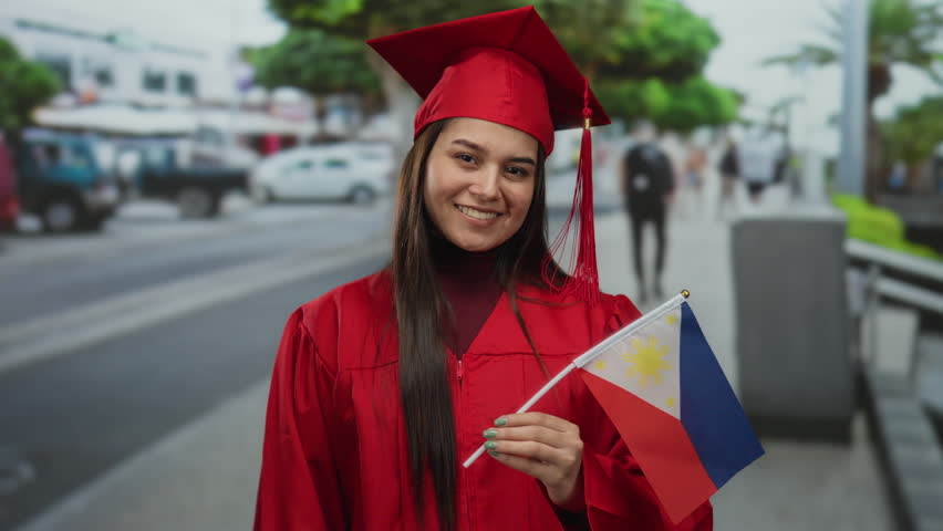 Young woman in red graduation gown proudly holding philippines flag on urban street, symbolizing achievement and national pride outdoors.