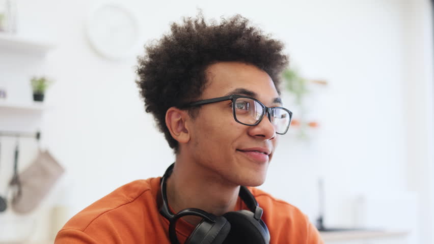 A young Black man with glasses smiles while looking up, with headphones around his neck.