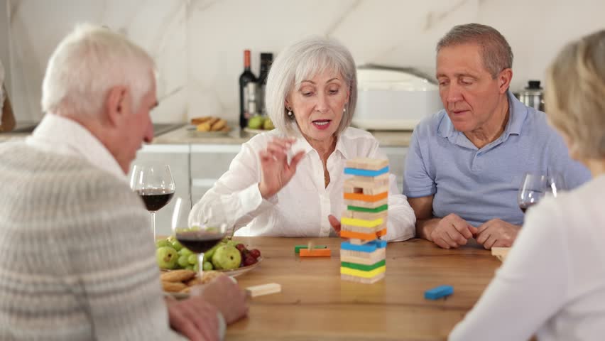 Group of four mature friends women and men is playing board game. Two senior married couples build tower with wooden blocks bricks 