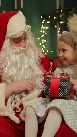 Santa Claus talks with a little girl while she holds a Christmas gift. Vertical video.