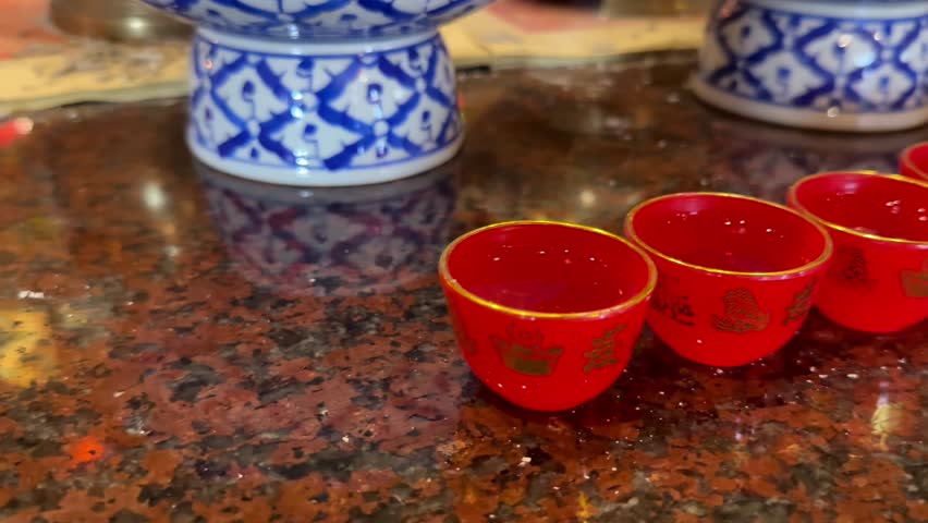 Horizontal close-up footage inside a Chinese Taoist temple showing a ceremonial offering table arranged according to traditional rituals. The camera begins with a row of small red offering cups filled with liquid, moving smoothly along them before revealing a glass oil lamp with a steady flame, symbolizing light, devotion and continuity. Nearby stand bronze incense vessels filled with incense sticks, some still burning while others have reduced to ash, representing prayers already offered and wishes still rising.

The scene continues across plates of ritual fruit offerings, including pineapple, mango, and green-skinned mandarins, placed as symbols of abundance, prosperity and gratitude. In front of each fruit plate sits a decorated bronze bowl filled with rice, into which incense sticks are placed, a common Taoist practice symbolizing nourishment, respect and communication with the spiritual realm.

Behind the offerings, richly dressed figurines of Taoist deities and guardian spirits are displayed against an ornate wall covered with traditional Chinese decorative patterns and calligraphy. The camera then gently tilts upward, revealing a bright ceiling adorned with intricate ornamental motifs and hanging Chinese lanterns, both traditional and modern in style. The calm, people-free atmosphere emphasizes spirituality, ritual symbolism and cultural heritage, making this footage suitable for documentaries, religious and cultural projects, Asian traditions, meditation visuals and Lunar New Year related content.