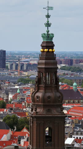 Aerial drone view of the ornate spire of Christiansborg Palace, crowned with golden spheres and intricate details, overlooking the cityscape of Copenhagen, Denmark. Vertical